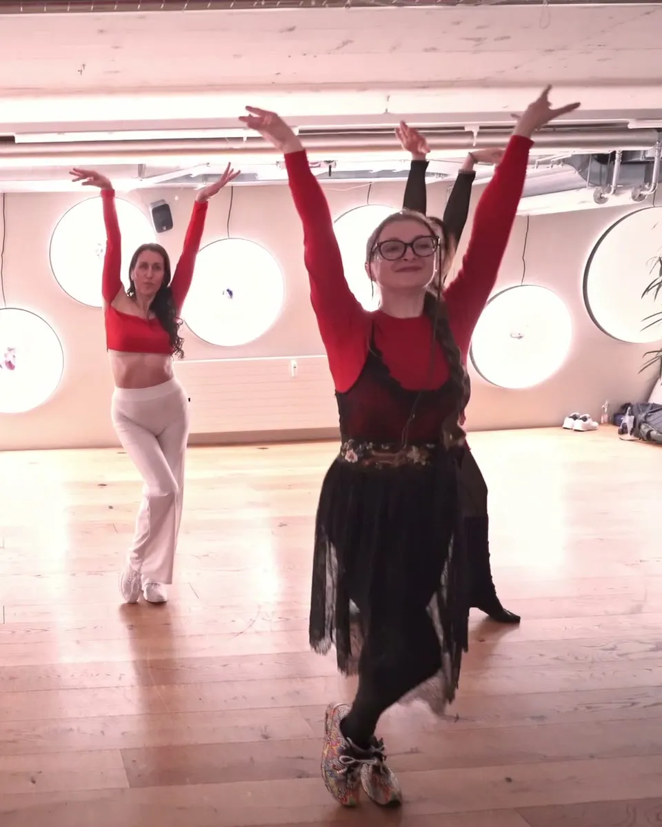 A dancer at the front of a Bachata Solo class, arms raised and smiling, the group behind her